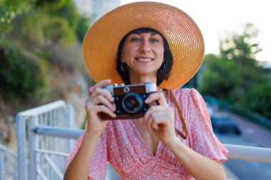 Pembe elbiseli ve şapkalı genç bir kadın şehirde kameraya çekiliyor. Analog bir kamerayla fotoğraf çekiyor. seyahat ve tatil.