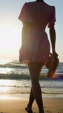 Dikey çekim. Gün batımında, elbise ve şapka giymiş bir kız sahilde yürüyor. Genç kız sahilde yaz tatilinin tadını çıkarıyor. Kız gün batımında sahilde yürüyor..