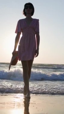 Dikey çekim. Gün batımında, elbise ve şapka giymiş bir kız sahilde yürüyor. Genç kız sahilde yaz tatilinin tadını çıkarıyor. Kız gün batımında sahilde yürüyor..