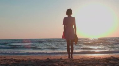Yazlık elbiseli ince kız deniz kıyısında duruyor ve gün batımını seyrediyor. Elbiseli mutlu kız ve elinde şapkasıyla kumsalda bir yaz tatilinin tadını çıkarıyor..