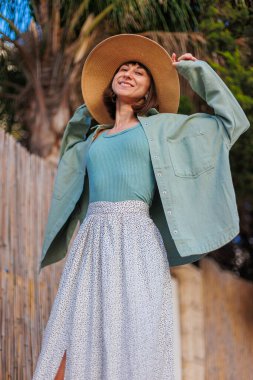 Genç bir esmer şehirde dururken mutlu bir şekilde gülümsüyor. Gömlekli, etekli ve şapkalı şık bir kız avlunun yanında duruyor. Yaz Portresi