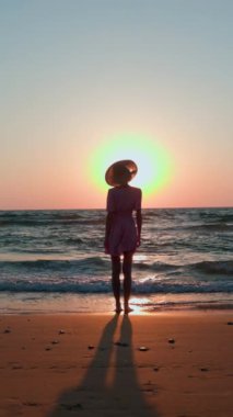 Dikey çekim. İnce bir kız deniz kıyısında duruyor ve gün batımını seyrediyor. Elbiseli ve şapkalı mutlu kız sahilde yaz tatilinin tadını çıkarıyor..