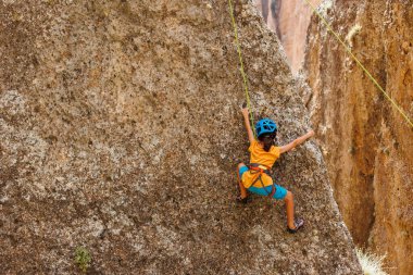 Yaz kampında spor. Bir çocuk yaz kampında kaya tırmanışı yapıyor. Dağcı çocuk. Güneşli bir günde doğada spor. Sevimli genç, kayaya bağlanmış bir şekilde tırmanıyor. Aktif tatil. Yaz tatili.