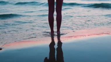 Kız sahilde duruyor ve sıcak dalgaların tadını çıkarıyor. Bacaklarını kapat. Gün batımında genç ve güzel bir kızın bacakları. Kumsalda dinlenmek. Yaz havası.
