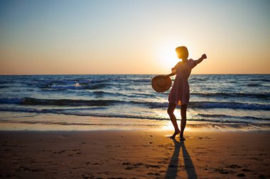 Sahilde yaz tatilinin tadını çıkaran, şapkalı ve elbiseli mutlu bir kızın silueti. Kız gün batımında sahilde yürüyor..