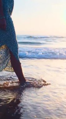 Dikey çekim. Sahilde elbiseli bir kız. Gün batımında sahilde uzun elbiseli genç bir kız yürüyor. Yaz havası.