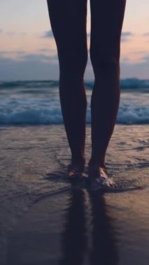 Dikey çekim. Kadın sahilde duruyor. Bacaklarını kapat. Deniz suyu gün batımında kızların ayaklarını yıkar. sakinlik ve kabullenme.