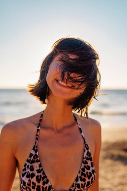 Yaz havası. Kumsalda güzel bir esmerin portresi. Genç mutlu kız gülümsüyor. Saçında rüzgar var. Saç yüzünü kapatır. Genç bir kız sahilde eğleniyor.