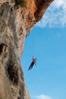 İple tırman. Genç bir adam kaya tırmanışı ile meşgul, yukarıdan bir atlet iniyor mavi gökyüzünün arka planında bir halat üzerinde.