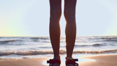 Sandaletli ince kız kumun üzerinde duruyor ve denize bakıyor. Yaz havası. Kızın bacakları yakın plan. Sahildeki kız..