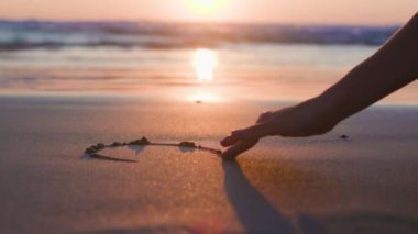 Sahilde gün batımında romantik bir yürüyüş. Yakın plan. Kız ıslak sahil kumu ve yapraklarına bir kalp işareti çiziyor. Romantik ruh hali.