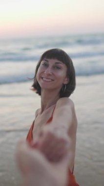 Dikey çekim. Beni takip edin. Mutlu genç kadın adamın elini tutuyor. Elbise giymiş güzel bir kız sahilde yürüyor, bir erkeğin elini tutuyor, romantik bir yürüyüş. Sahilde yürü..
