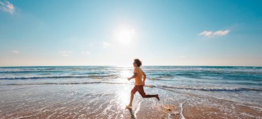 Adam deniz kıyısında çıplak ayakla koşuyor. Güçlü koşucu, hızlı koşu ve yaz antrenmanı. Genç bir erkek koşucu şafakta boş bir plaj boyunca koşar. Denize yakın koş..