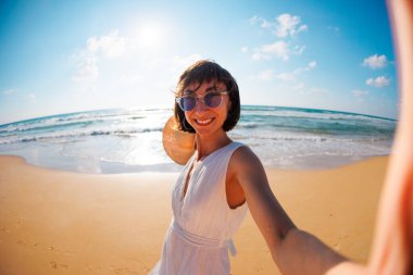 Sevimli, şaşkın, güzel bir kızın sahilde yürürken ve kameraya selfie çekerken çekilmiş fotoğrafı. Kız selfie çekiyor..