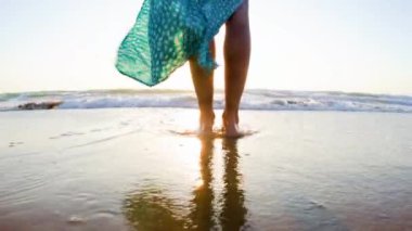 Sahilde çıplak ayaklı, güzel elbiseli bir kız. Elbiseli bir kadın kumlu bir sahilde gün batımını seyrediyor. bacaklar kapalı..