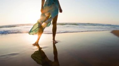 Sahilde çıplak ayaklı, güzel elbiseli bir kız. Elbiseli bir kadın kumlu bir sahilde gün batımını seyrediyor. bacaklar kapalı..