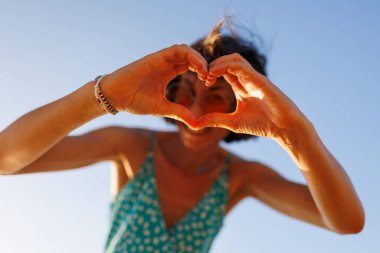 Genç kız kalp yapar. Aşkın şeklinin bir sembolü. Turuncu güneş gözlüklü neşeli kadın elleriyle kalp şekli yapıyor. Kız sahilde eğleniyor..
