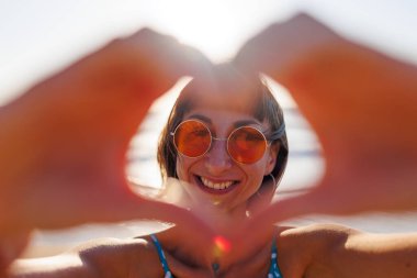 Genç bir kadının kalp şeklini oluşturan ellerine yakın çekim. Kameraya bakarken parmaklarıyla kalp yapan güzel bir kadının portresi. Gülümseyen genç kız gün batımında aşk işareti yapıyor..