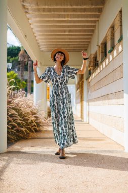 Şapka takan güzel bir bayanın tam boy fotoğrafı. Sokakta yürümek, güneşli hava, hafta sonu. Elbiseli ve şapkalı genç bir kız sokakta yürüyor..