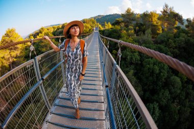 Sırt çantalı bir kız. Bir nehrin üzerindeki asma köprüde yürüyen genç bir kadının açık hava portresi. Seyahat ve macera