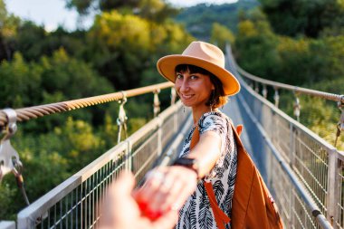 Sırt çantalı bir kız. Bir nehrin üzerindeki asma köprüde yürüyen genç bir kadının açık hava portresi. Kız döndü ve sanki beni takip ediyormuş gibi kameraya elini uzattı. Seyahat ve macera