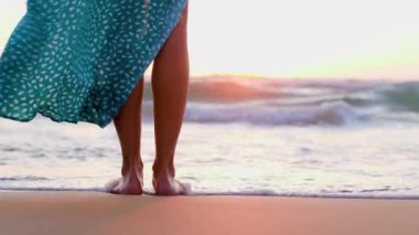 Sahilde çıplak ayaklı, güzel elbiseli bir kız. Elbiseli bir kadın kumlu bir sahilde gün batımını seyrediyor. bacaklar kapalı..