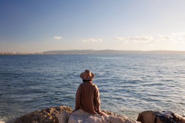 Arkadan bak. Paltolu ve şapkalı genç bir kız yüksek bir taşın üzerinde oturuyor ve mavi denizin ve gökyüzünün güzel manzarasına bakıyor. Kıyı şeridi.