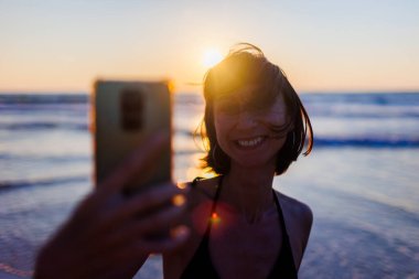 Akıllı telefonlu güzel bir genç gün batımında deniz sahilinde yürüyor, selfie çekiyor. Güneşin doğuşunda sahilde gülümseyen telefon kamerası. Kadın selfie çekiyor.