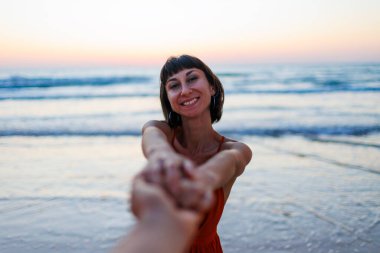 Kız sahilde eğleniyor. Elbise giymiş güzel bir kız sahilde yürüyor, bir erkeğin elini tutuyor, romantik bir yürüyüş. Sahil boyunca yürü..