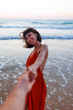 Kız sahilde eğleniyor. Elbise giymiş güzel bir kız sahilde yürüyor, bir erkeğin elini tutuyor, romantik bir yürüyüş. Sahil boyunca yürü..