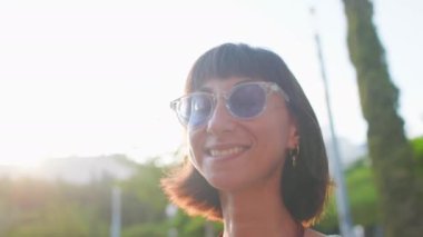 Şehirdeki genç bir kadın. Parlak güneşli bir günde, şık kıyafetler ve güneş gözlükleriyle sevimli bir gülümsemesi olan bir kadın. Kız şehirde gezmekten zevk alıyor..