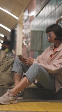 Dikey çekim. İstasyonda tren bekleyen ve telefon kullanan genç bir kadın. Telefona yakın çekim. Programa bak. Metroda elinde telefonla bir kız..