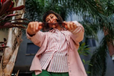 Geniş gülümseyen bir kızın portresi. Kız pembe gömlek ve yeşil pantolon giyiyor. Kentsel giyim tarzı. Zarif kaygısız kız parkta eğleniyor. Duygusal esmer model tatilinin tadını çıkarıyor..