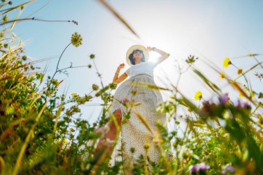 Yaz havası. Mavi gökyüzü ve çiçeklerle dolu bir arka planda poz veren genç ve neşeli bir kız. Çiçeklerin arasında mutlu bir kız ve bahar doğası dışarıda dinleniyor. bahar modası.