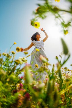Yaz havası. Mavi gökyüzü ve çiçeklerle dolu bir arka planda poz veren genç ve neşeli bir kız. Güzel bir kız çiçekler arasında dans eder. Çiçeklerin arasında dans eden kız. bahar modası.
