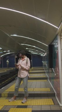 Dikey çekim. Kız treni bekliyor. Metrodaki kız. Metroda akıllı telefon kullanan, şık elbiseli, sırt çantalı genç bir kız. moda ve tarz.