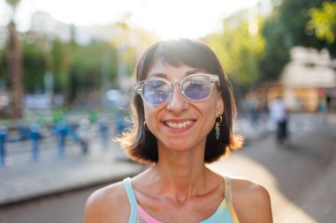 Neşeli, neşeli, genç bir kadının portresi. Hoş bir gülümsemesi var. Şık kıyafetler giyiyor ve güneşli bir günde açık havada güneş gözlüğü takıyor. Kız şehirde yürümekten zevk alıyor..