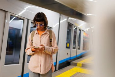 Sırt çantalı genç bir kız metroda akıllı telefon kullanıyor. Kız tren bekliyor. Metrodaki kız..