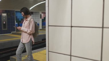 Genç bir kadın metro istasyonunda duruyor ve cep telefonuna bakıyor. Bir kadın metro istasyonunda cep telefonu kullanıyor. Kız programa bakıyor..