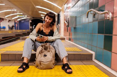 Young woman waiting for a train at the station. wait for a train. girl in the subway. Young girl in stylish clothes with a backpack in the subway. fashion and style. clipart