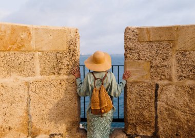 Sırt çantalı şık bir kız eski şehrin caddesinde duruyor ve denize bakıyor. Maceralar ve seyahat.