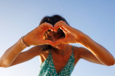 Genç bir kadının kalp şeklini oluşturan ellerine yakın çekim. Kameraya bakarken parmaklarıyla kalp yapan güzel bir kadının portresi. Gülümseyen genç kız gün batımında aşk işareti yapıyor..