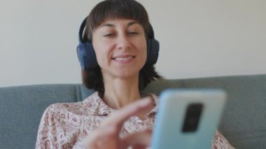 Gülümseyen kız evde dinleniyor, müzik dinliyor, akıllı telefon kullanıyor. Oturma odasında, evdeki kanepede, güzel sesleri dinleyen genç bir kadın..