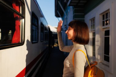 Genç gezgin platformda duruyor ve trendeki birine el sallayarak veda ediyor. Gitmeden önce hoşça kal de. seyahat ve macera.