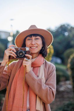 Şık bir görünüş, kameralı genç bir kadın zarif bir şapka ve ceket giyiyor. Hipster bir turist kadın elinde kamerayla fotoğraf çekiyor. film kamerası.