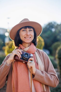 Şık bir görünüş, kameralı genç bir kadın zarif bir şapka ve ceket giyiyor. Hipster bir turist kadın elinde kamerayla fotoğraf çekiyor. film kamerası.