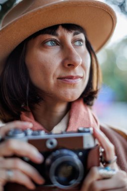 Şık bir görünüş, kameralı genç bir kadın zarif bir şapka ve ceket giyiyor. Hipster bir turist kadın elinde kamerayla fotoğraf çekiyor. film kamerası.