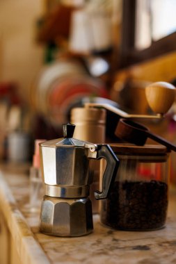 Evdeki masada Moka pot var. Kahve yapma konsepti. Moka demliği, fasulye spreyi, kahve öğütücü, mutfak masasının üzerinde duran bir kavanoz kahve çekirdeği. Kahve yapmak için gerekli malzemeler.