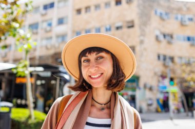 Güneşli bir havada şehrin arka planına karşı şapkalı neşeli beyaz bir kadının portresi. İlginç yerlere seyahat et. Kameraya gülümseyen şapkalı şık bir kızın fotoğrafı..