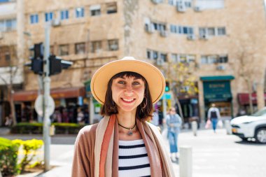 Güneşli bir havada şehrin arka planına karşı şapkalı neşeli beyaz bir kadının yakın plan portresi. Hafta sonu gezisi. Kameraya gülümseyen şapkalı şık bir kızın fotoğrafı..